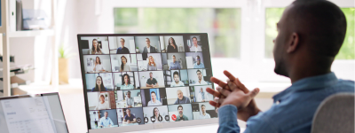 A man sitting at a desk with lots of people on a virtual meeting on the screen in front of him.